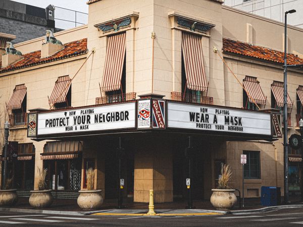 Photograph of old movie theatre. Two marquee signs say: NOW PLAYING, PROTECT YOUR NEIGHBOR, WEAR A MASK.